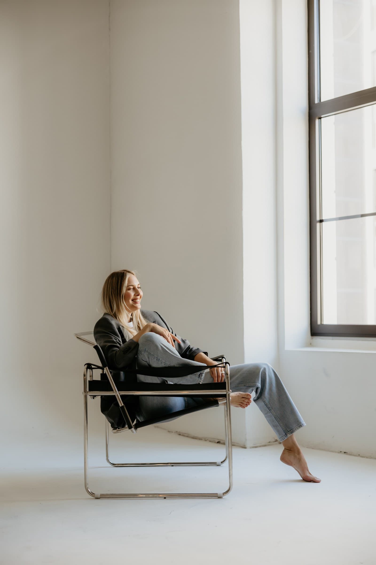 Lea sits in a low chair by a tall window, light falling across an empty wall.