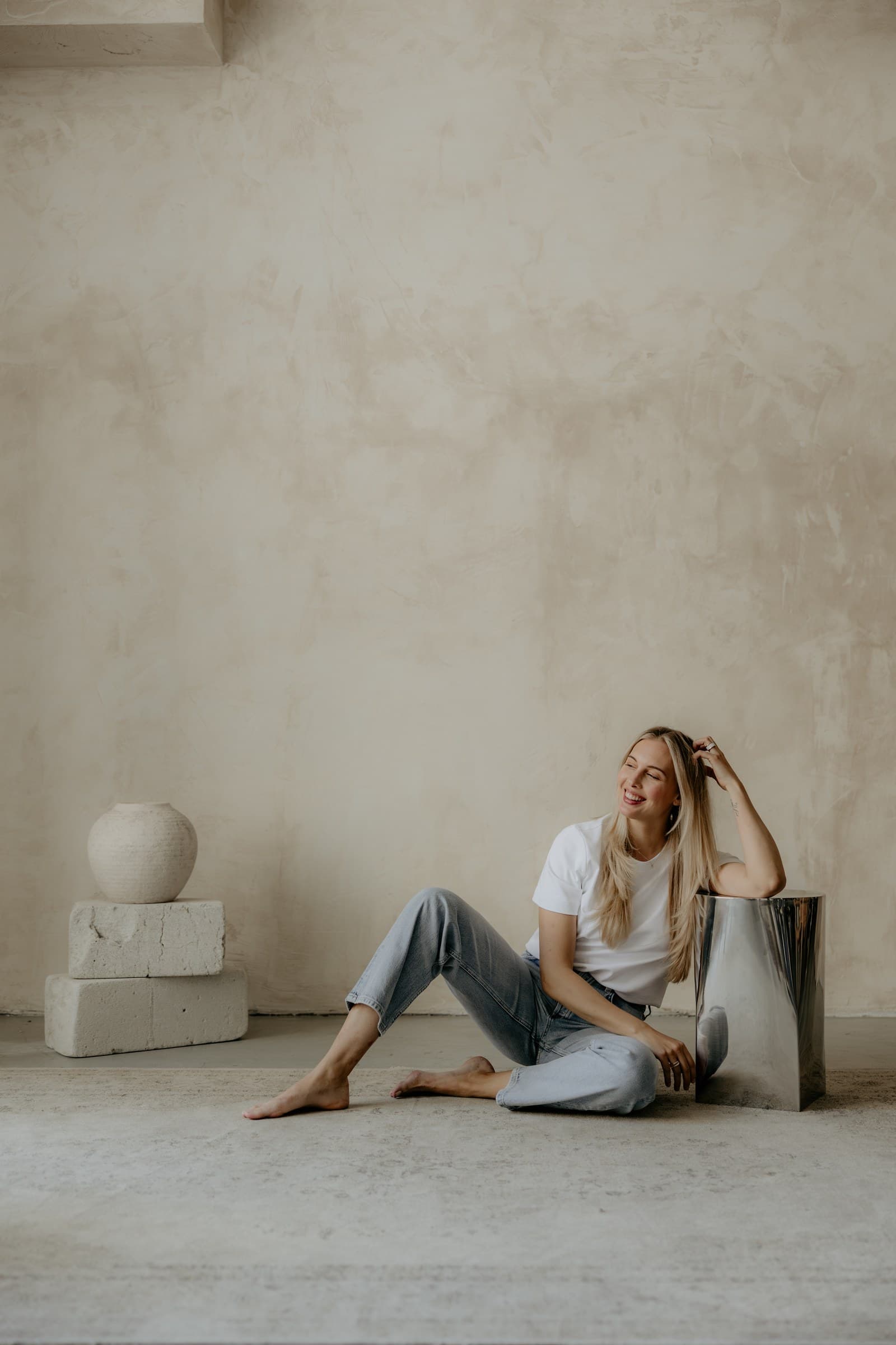 Lea sits on the floor by a plaster wall, beside a clay vase on stone, in warm and quiet daylight.